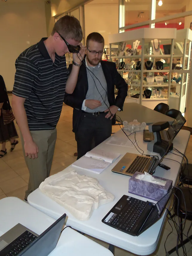 Two visitors at a table of laptops and plaster objects, one wearing an augmented-reality headset.