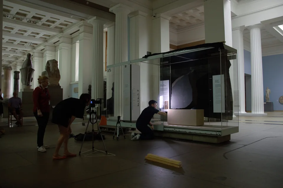 The Rosetta Stone in its glass case in a British Museum gallery while a team sets up scanning equipment on the floor.