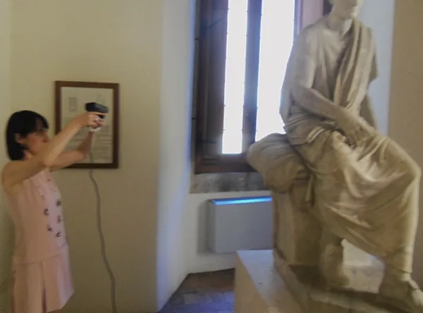 A visitor holding a handheld depth sensor to scan a seated marble statue in a museum room.