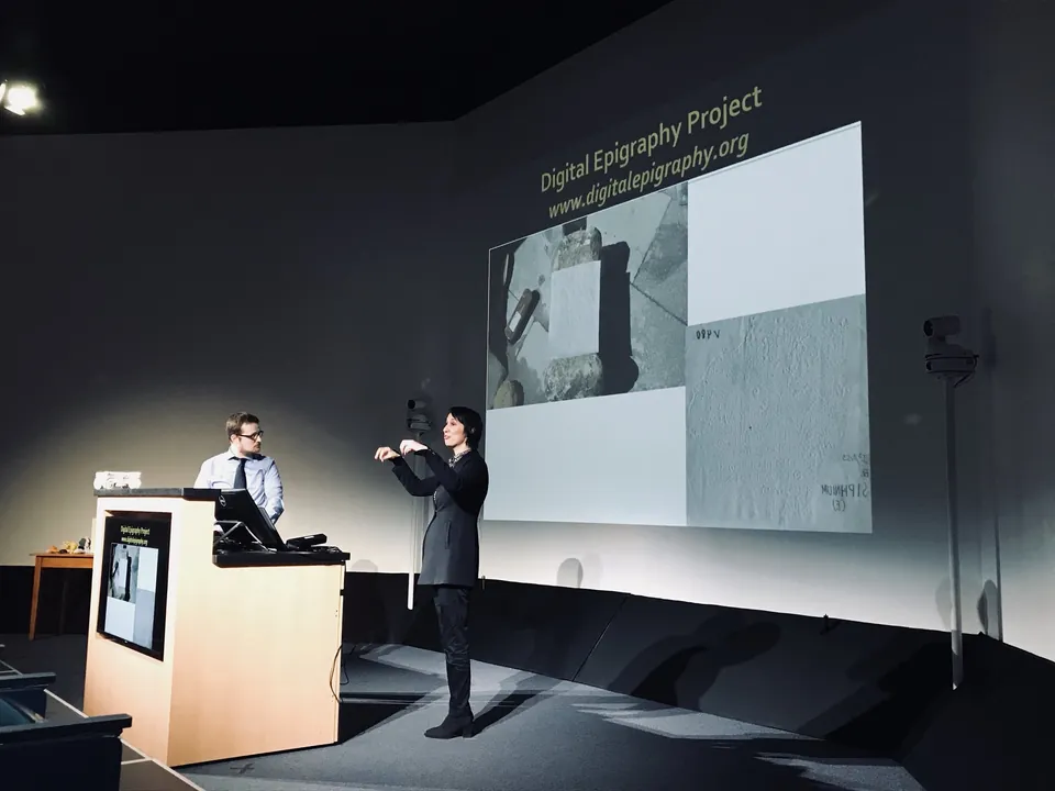 Two presenters on a stage beside a screen reading “Digital Epigraphy Project”.
