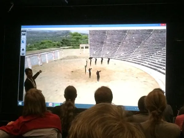 An audience watching a presentation showing an ancient theatre with figures standing in its orchestra.