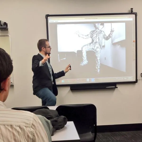 A speaker presenting in a classroom beside a screen showing a 3D scan of a statue.