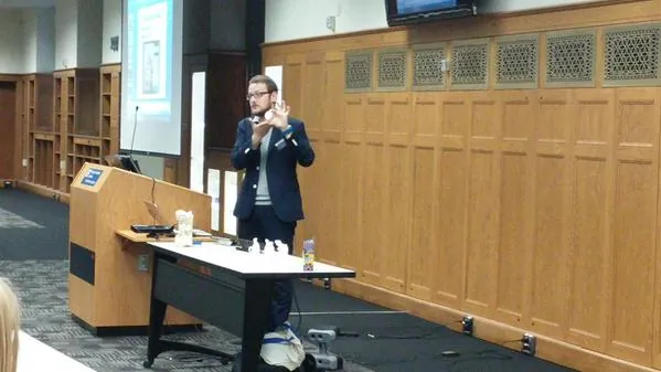A speaker presenting at a lectern in a wood-paneled lecture hall.